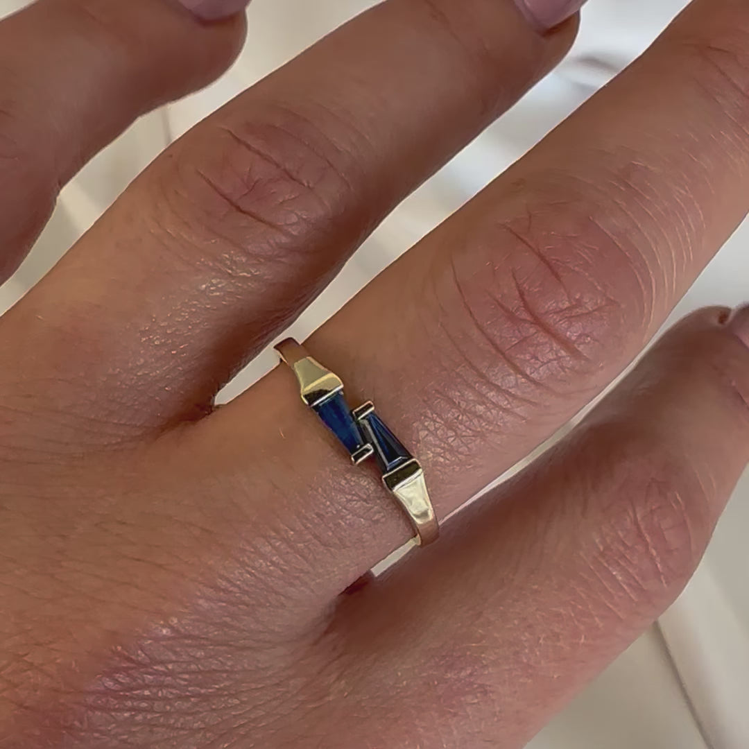 a gold ring with two tapered baguette blue sapphires displayed on a women's hand