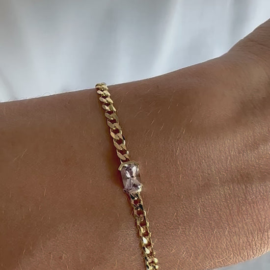 A gold bracelets with a curb chain design one featuring an emerald cut natural pink sapphire displayed on a person's wrist.