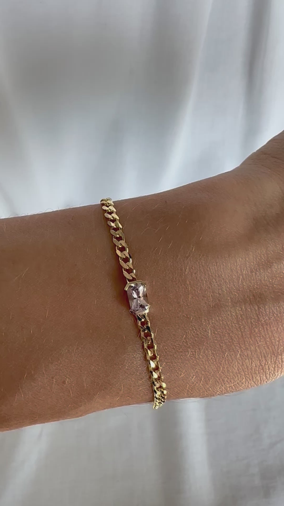 A gold bracelets with a curb chain design one featuring an emerald cut natural pink sapphire displayed on a person's wrist.
