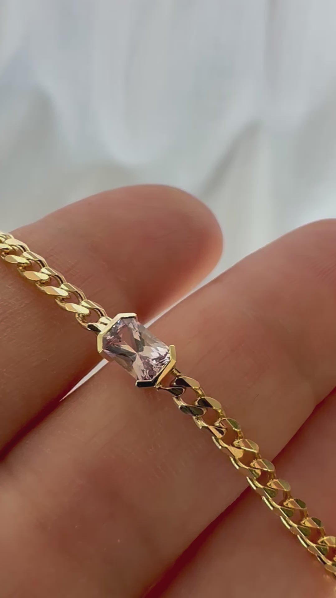A gold bracelets with a curb chain design one featuring an emerald cut natural pink sapphire displayed in person's hand