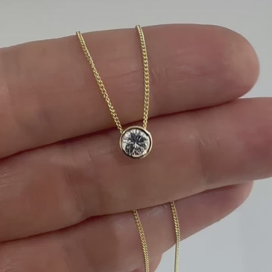 Round diamond bezel pendant on a fine 14K yellow gold chain, held between the fingers.