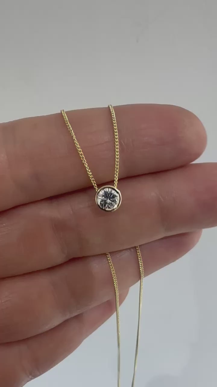 Round diamond bezel pendant on a fine 14K yellow gold chain, held between the fingers.