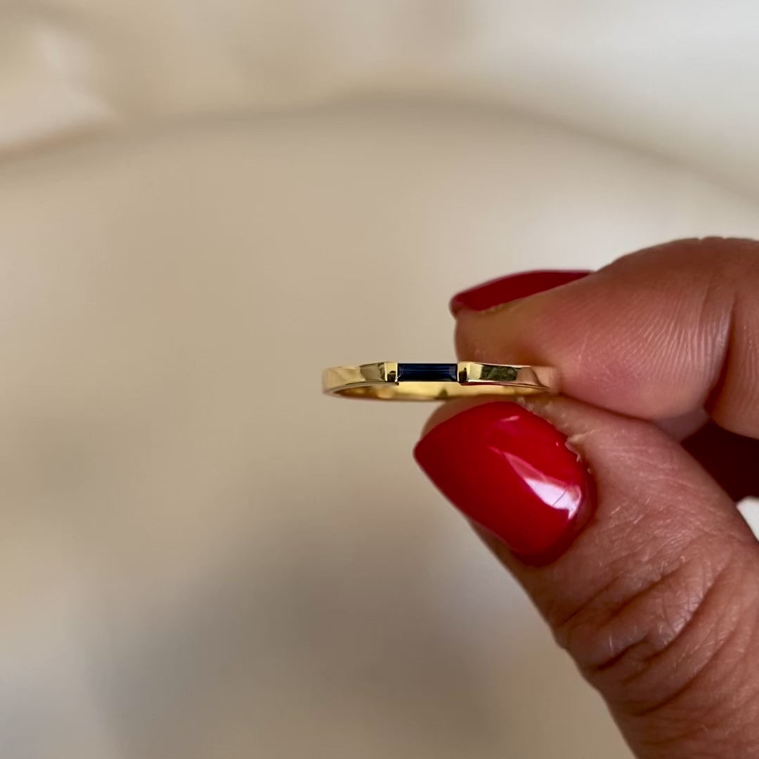 A minimalist style ring with a needle baguette cut blue sapphire set in a 14K yellow gold band, shown in womens hand against a white background.