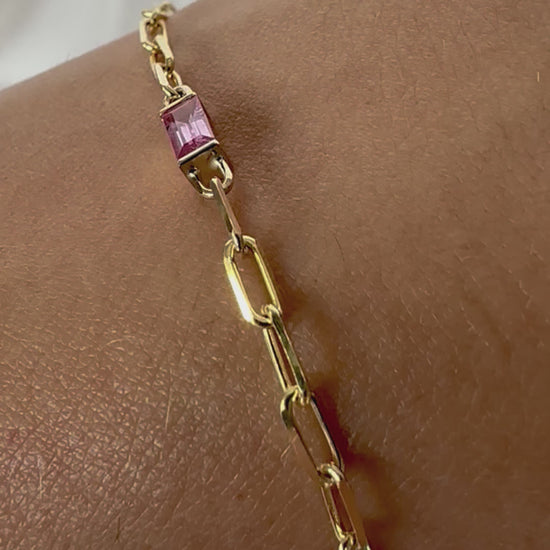 A gold bracelet with a pink sapphire baguette cut paper clip chain, displayed on a person's wrist.
