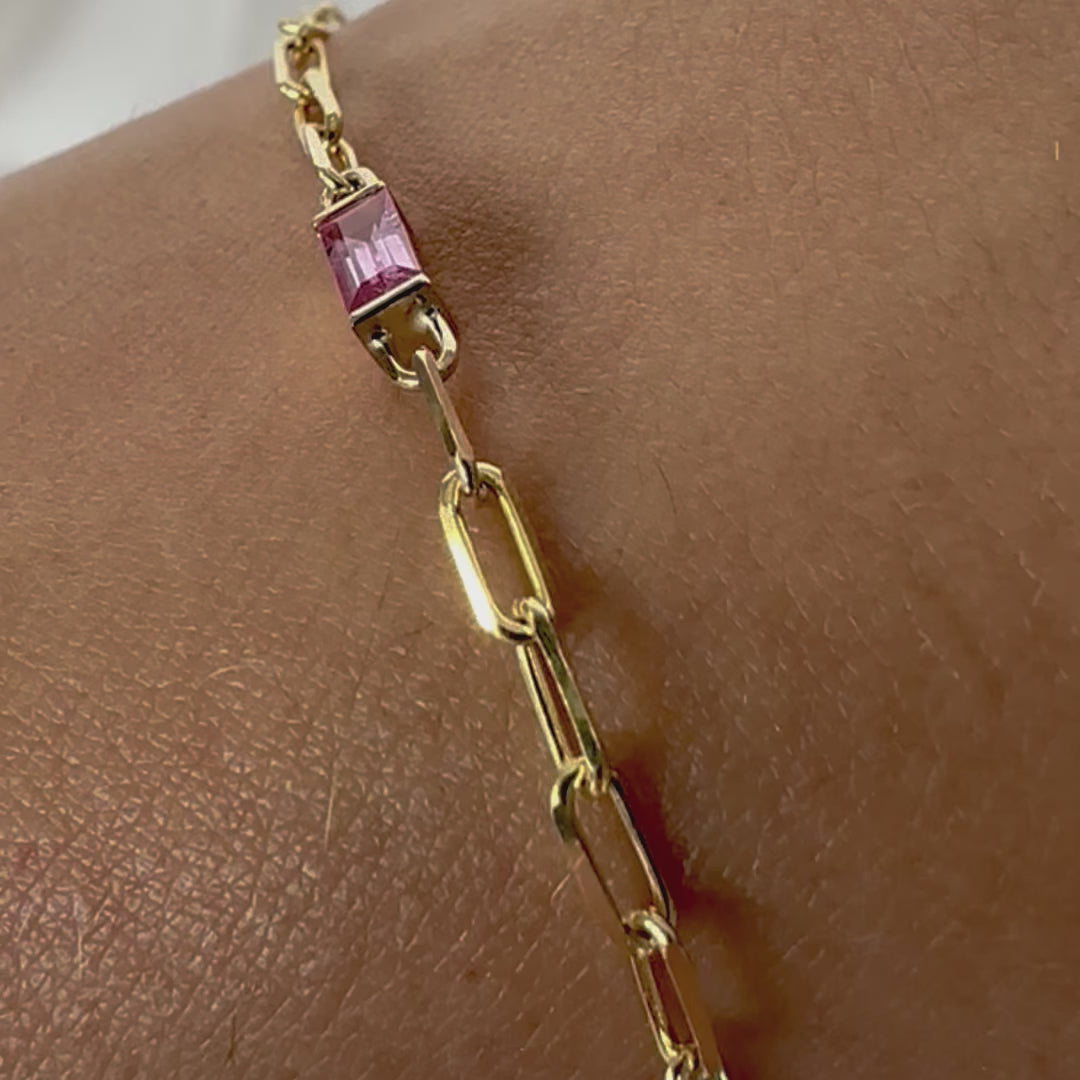 A gold bracelet with a pink sapphire baguette cut paper clip chain, displayed on a person's wrist.