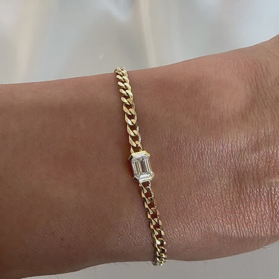 A gold bracelet with a curb chain design featuring an emerald cut diamond solitaire, displayed on a person's wrist.