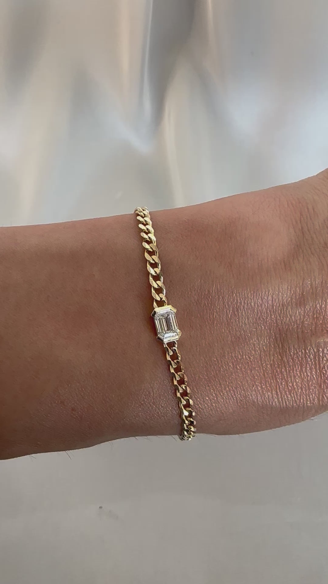 A gold bracelet with a curb chain design featuring an emerald cut diamond solitaire, displayed on a person's wrist.