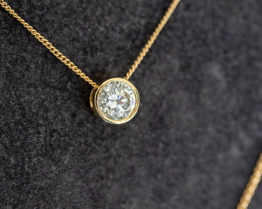 Round brilliant diamond in a 14K yellow gold bezel pendant, close-up on dark grey felt.