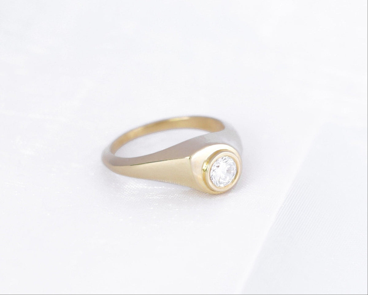 Drop-shaped 14K yellow gold signet ring with a bezel-set round diamond, angled view on white fabric