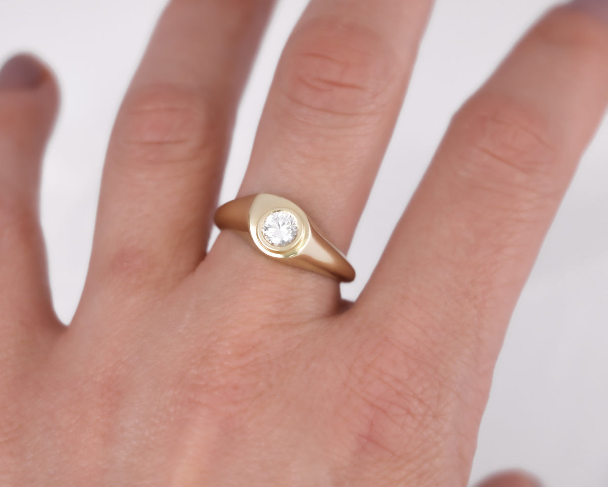 Drop-shaped 14K yellow gold signet ring with a round diamond in a bezel, worn on the ring finger
