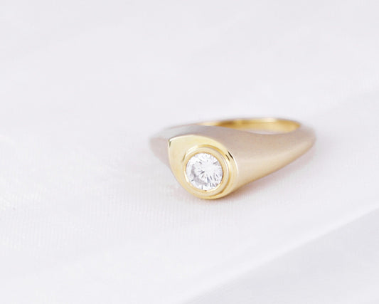 Round diamond in a bezel on a drop-shaped 14K yellow gold signet ring, three-quarter view on white fabric