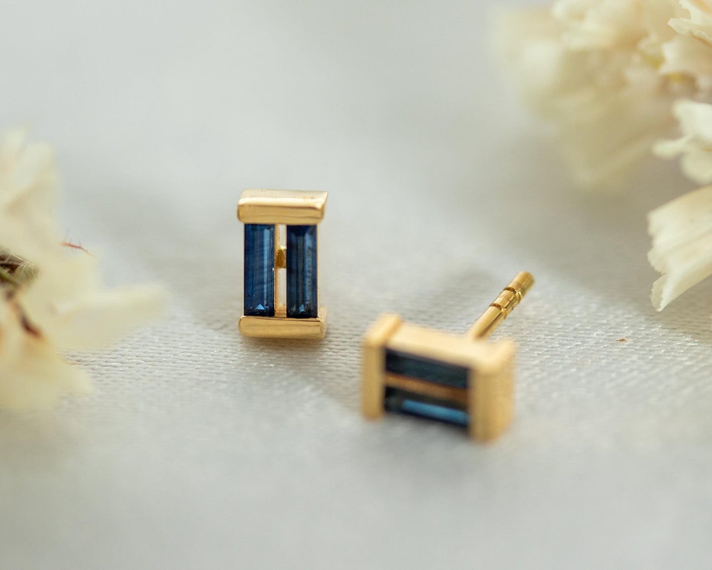Pair of gold baguette blue sapphire stud earrings on a white surface with dried flowers
