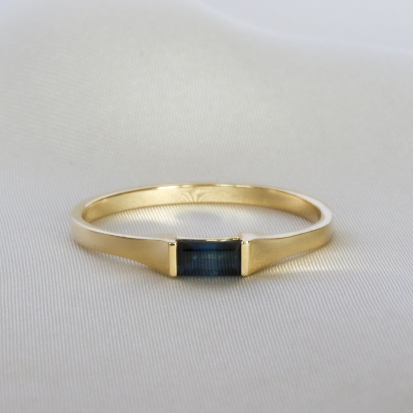 A sleek baguette cut blue sapphire ring in 14K yellow gold, displayed against a satin background.