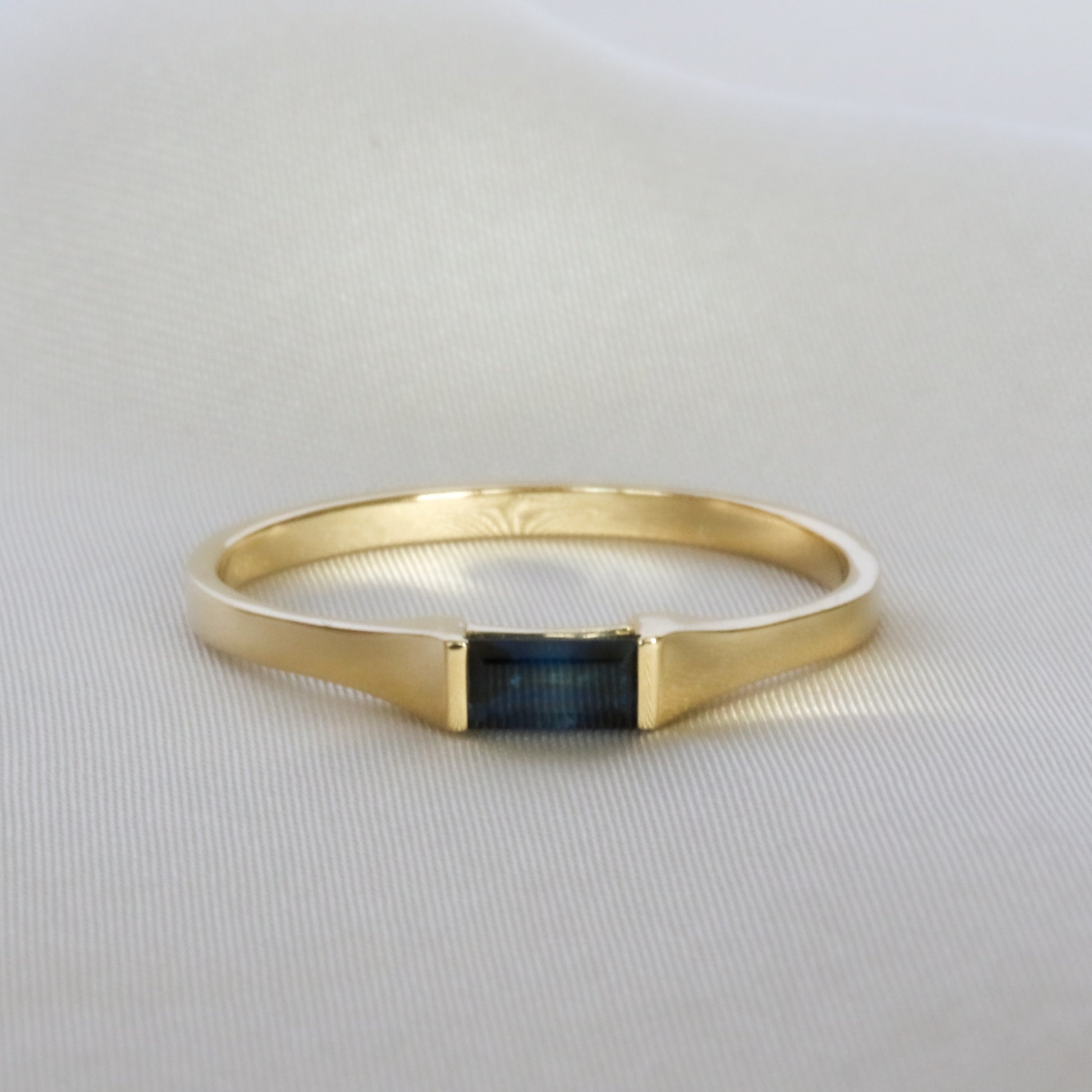A sleek baguette cut blue sapphire ring in 14K yellow gold, displayed against a satin background.