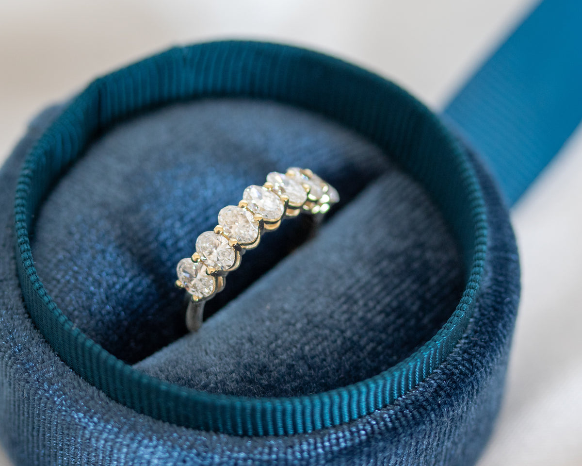 Seven-stone oval-cut diamond ring resting in a teal jewelry box with ribbon