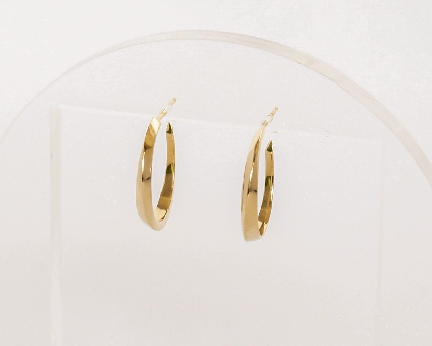 Small 14K gold twist hoop earrings with geometric sculpted design, minimalist modern jewelry displayed on clear acrylic stand with soft lighting.