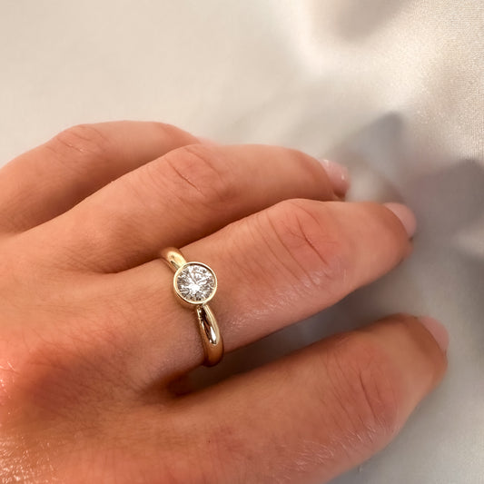 A hand wearing a solitaire bezel diamond ring with a gold band.