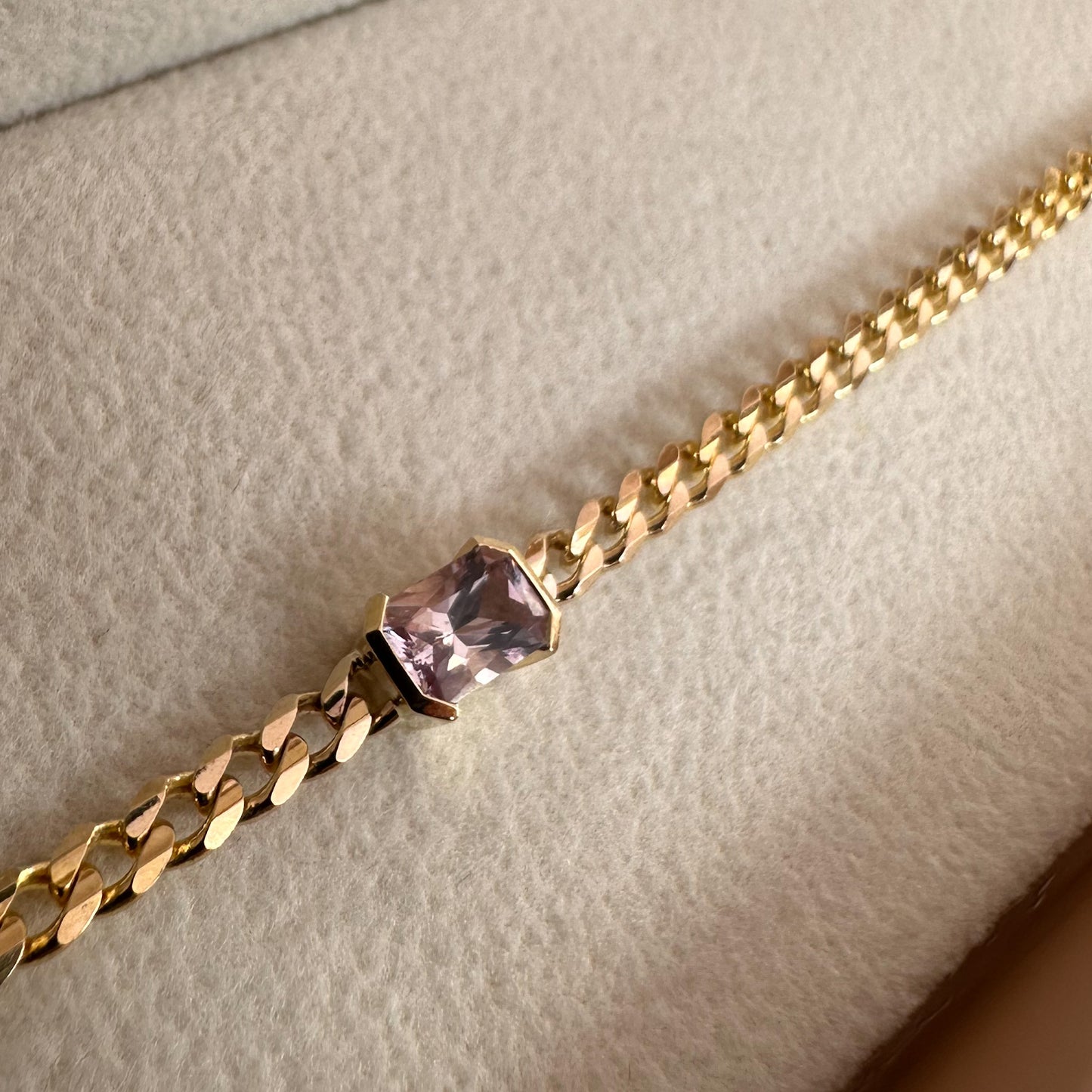 A gold bracelets with a curb chain design one featuring an emerald cut natural pink sapphire displayed on white textured background