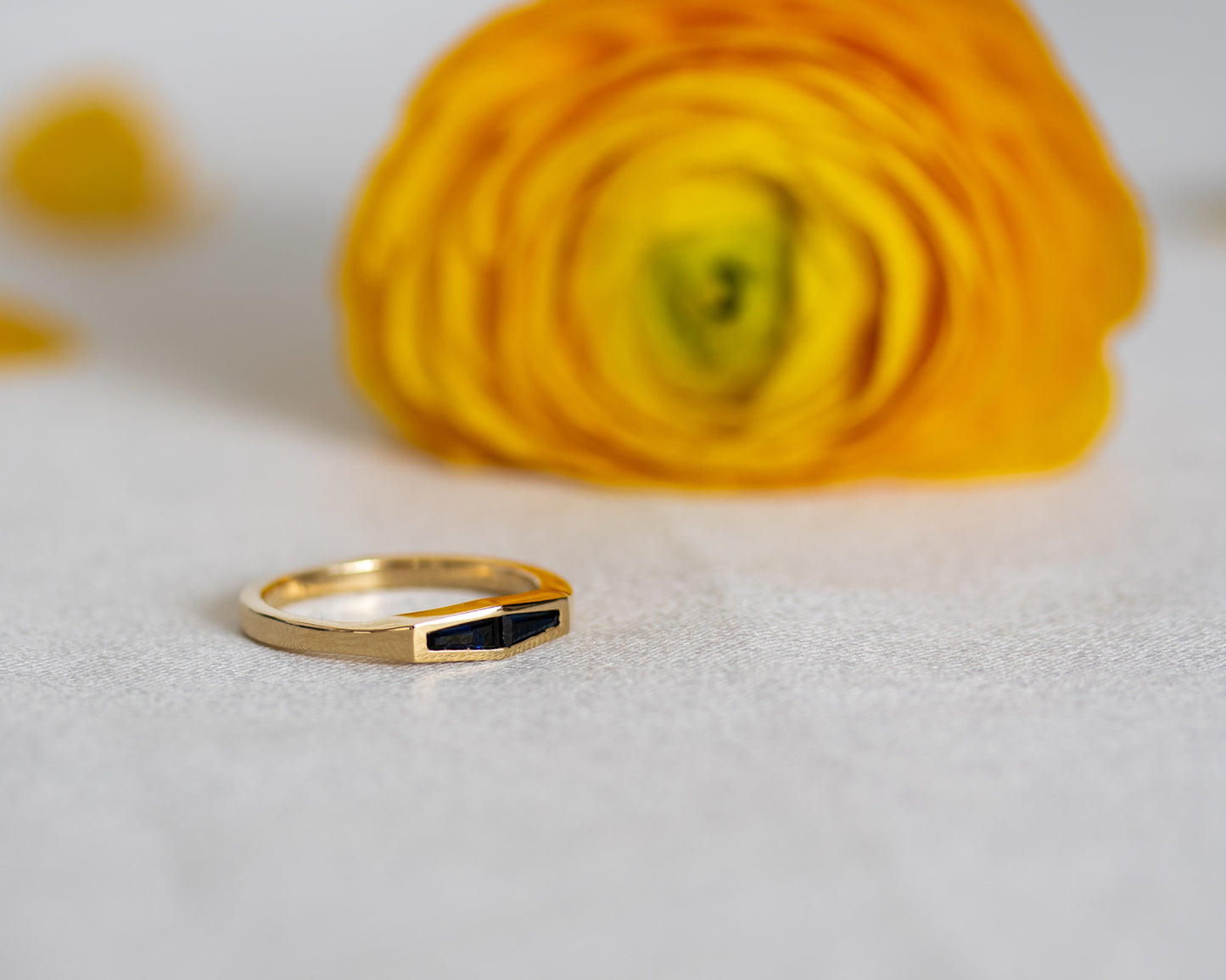 Two-stone blue sapphire ring in 14K yellow gold, styled beside a yellow flower.