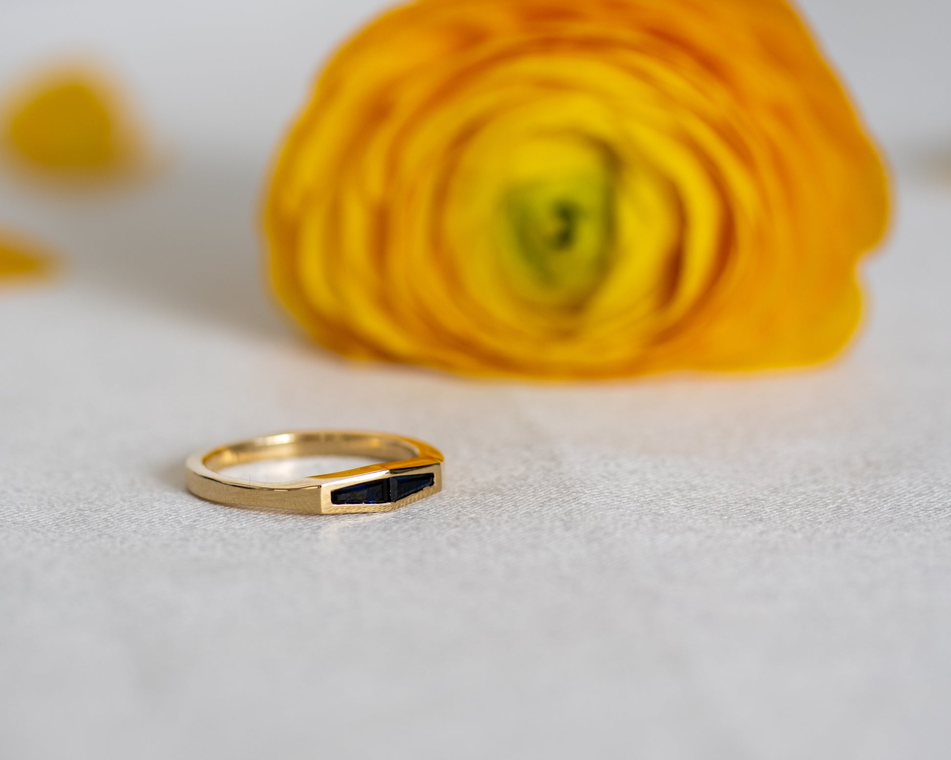 Two-stone blue sapphire ring in 14K yellow gold, styled beside a yellow flower.