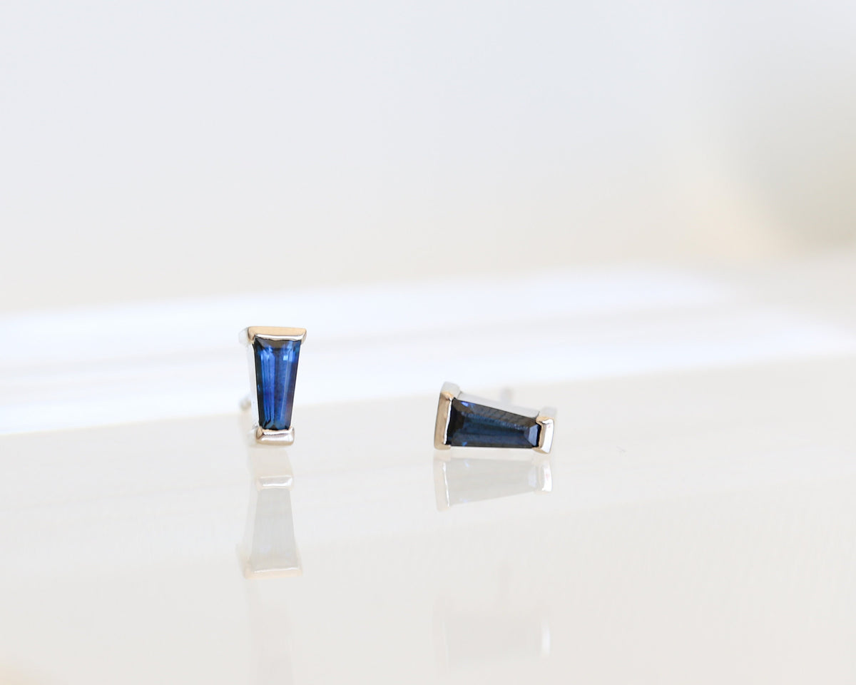 Pair of blue tapered baguette sapphire studs in white gold, one upright and one on its side, on white
