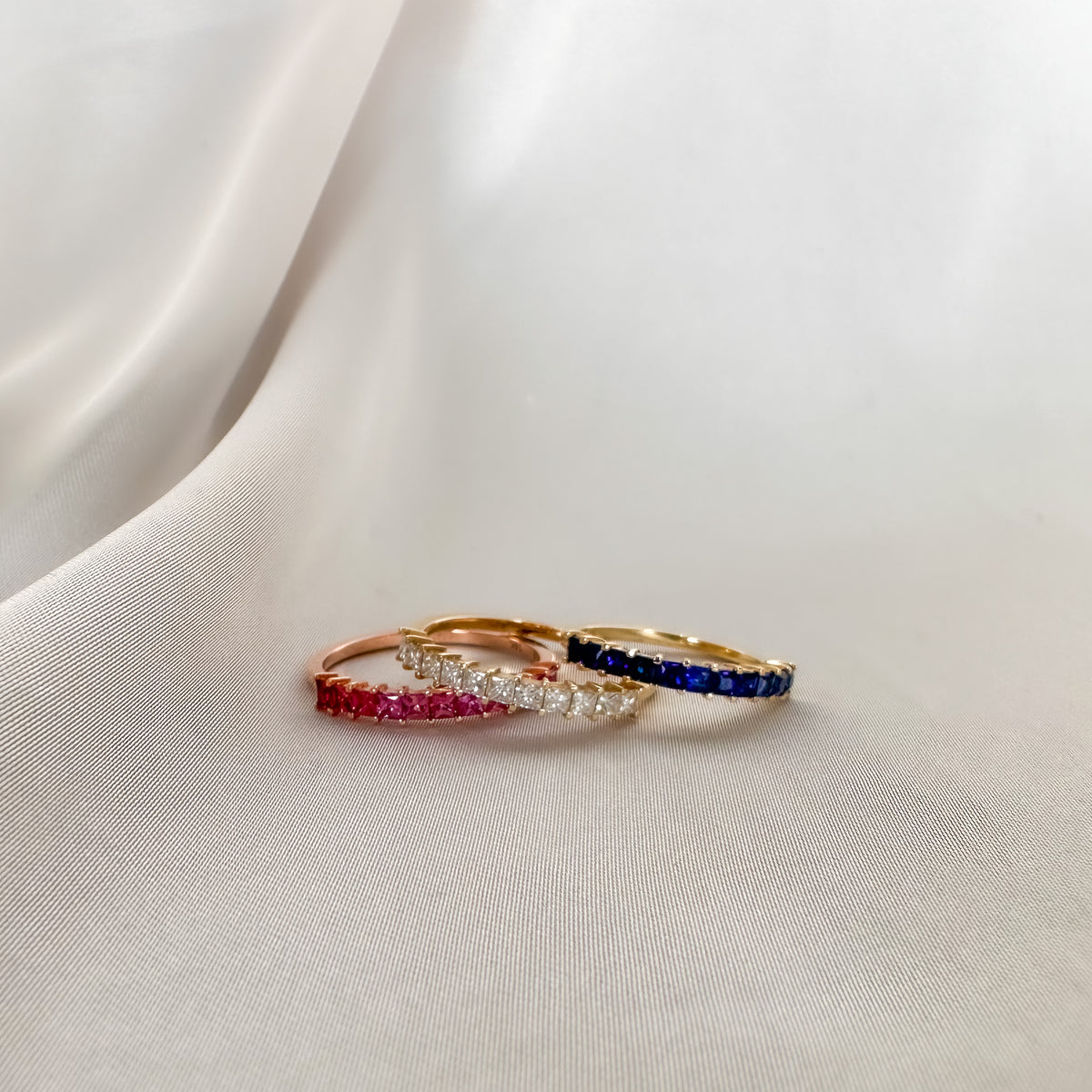 Three princess cut half eternity bands in 14K gold with diamonds, pink sapphires and blue sapphires on white fabric