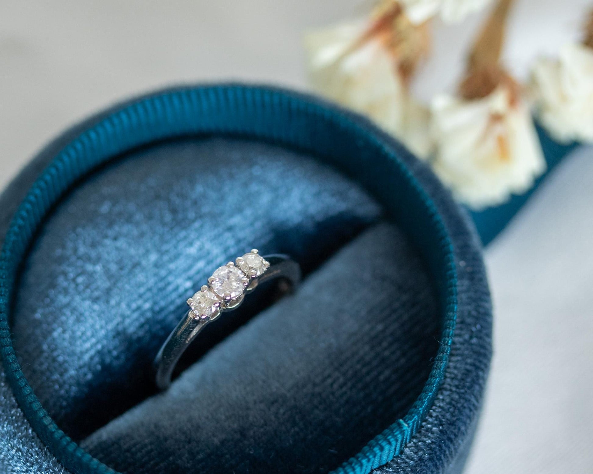 Three-stone diamond ring in 14K white gold, classic trilogy engagement design displayed in blue velvet box with soft floral background