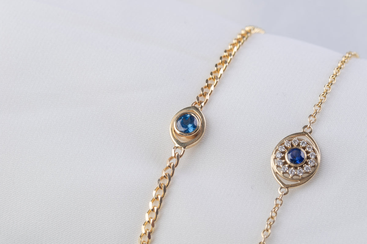 Two 14K yellow gold evil eye bracelets with blue sapphires, with and without diamonds, on a light background