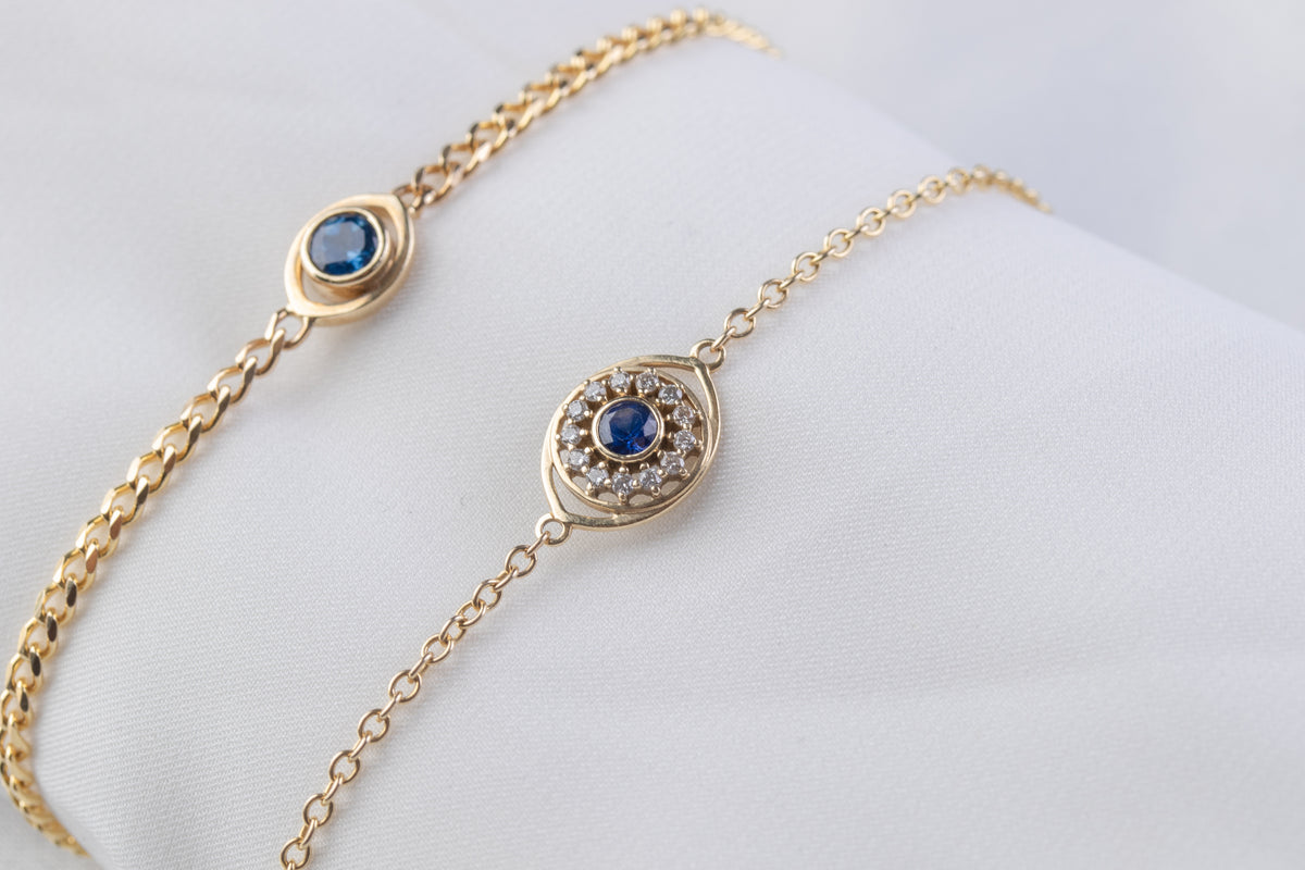 Two 14K yellow gold evil eye bracelets with blue sapphires, with and without a diamond halo, on a white background
