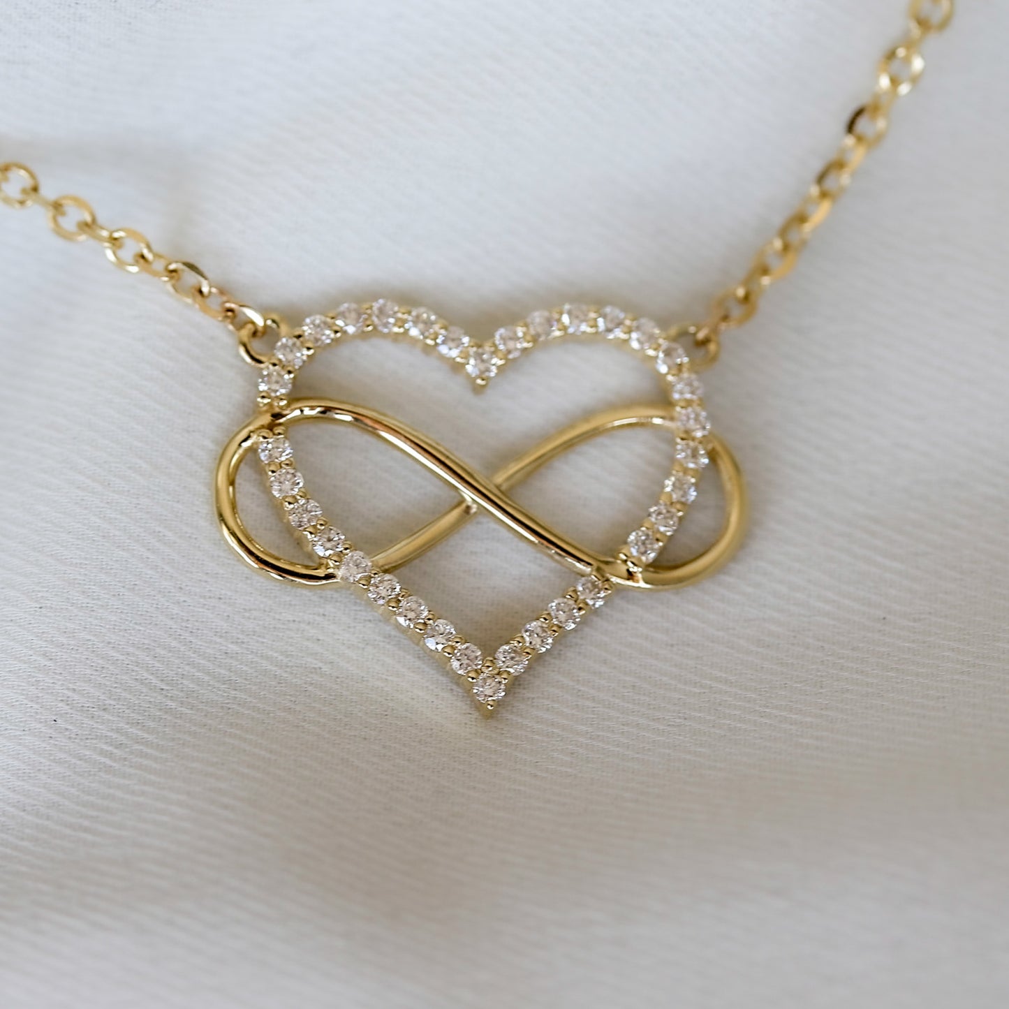 A heart-shaped pendant necklace with a diamonds and gold infinity symbol, attached to a yellow gold chain, displayed on white background