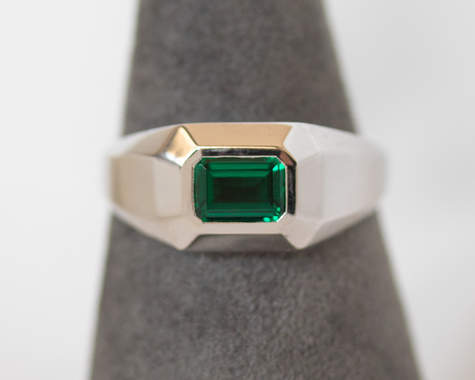 Men’s white gold signet ring with emerald-cut green Chatham emerald on a gray display stand