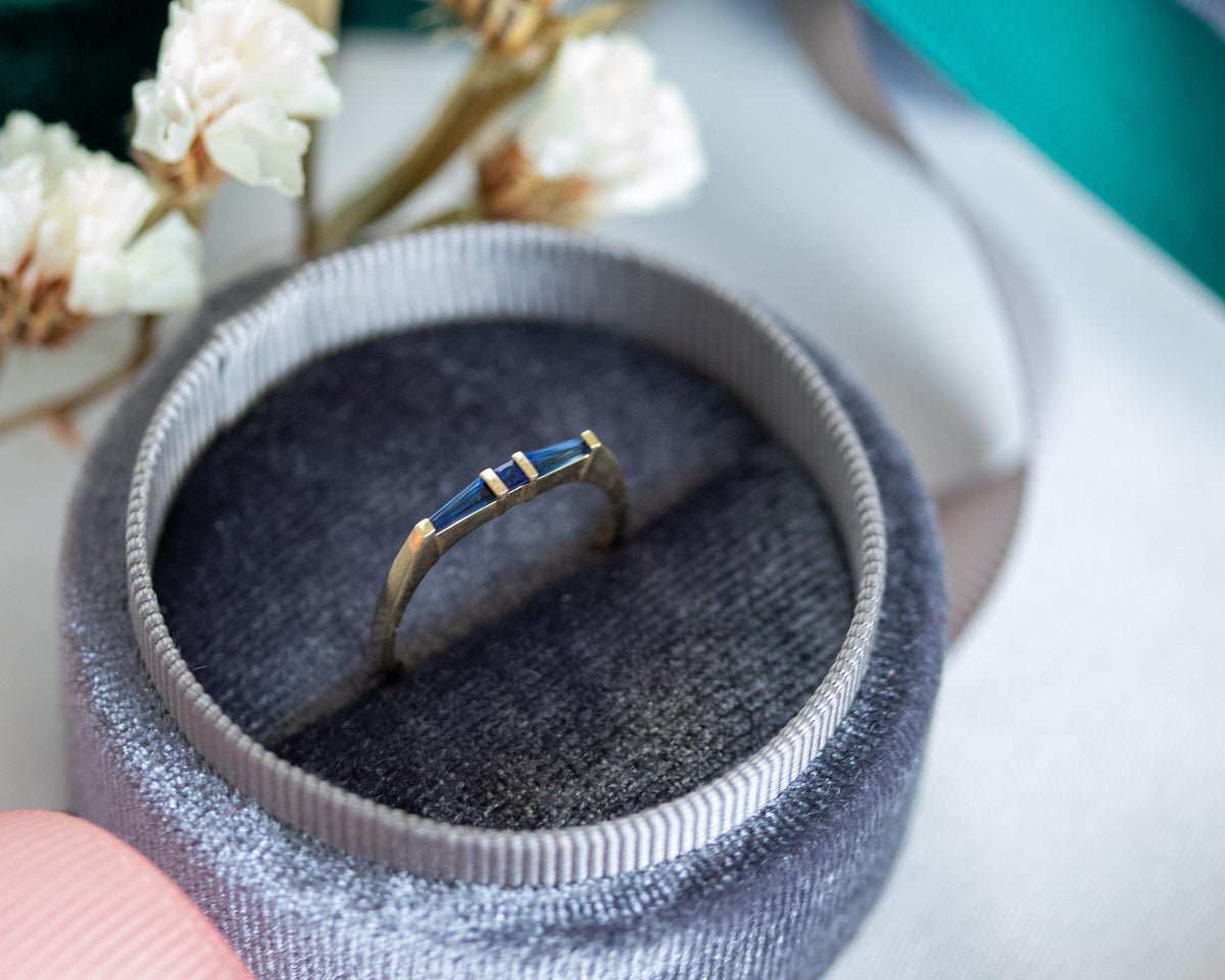 Three-stone blue sapphire ring in yellow gold in an open velvet ring box, with dried flowers and ribbon