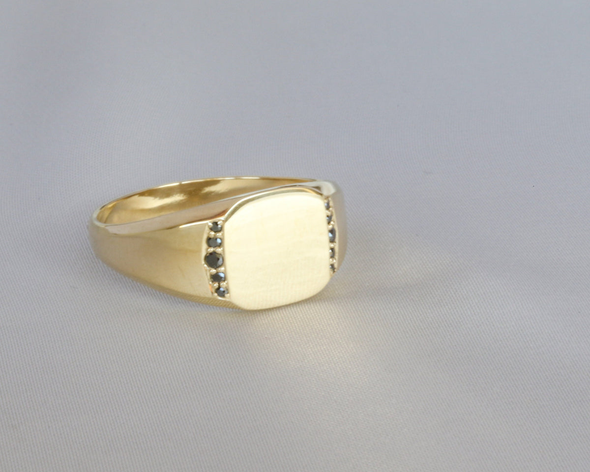 Yellow gold men's signet ring with black diamonds set flush below the flat top, on a light gray background