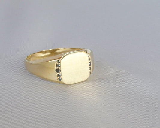 Yellow gold men's signet ring with black diamonds set flush below the flat top, on a light gray background