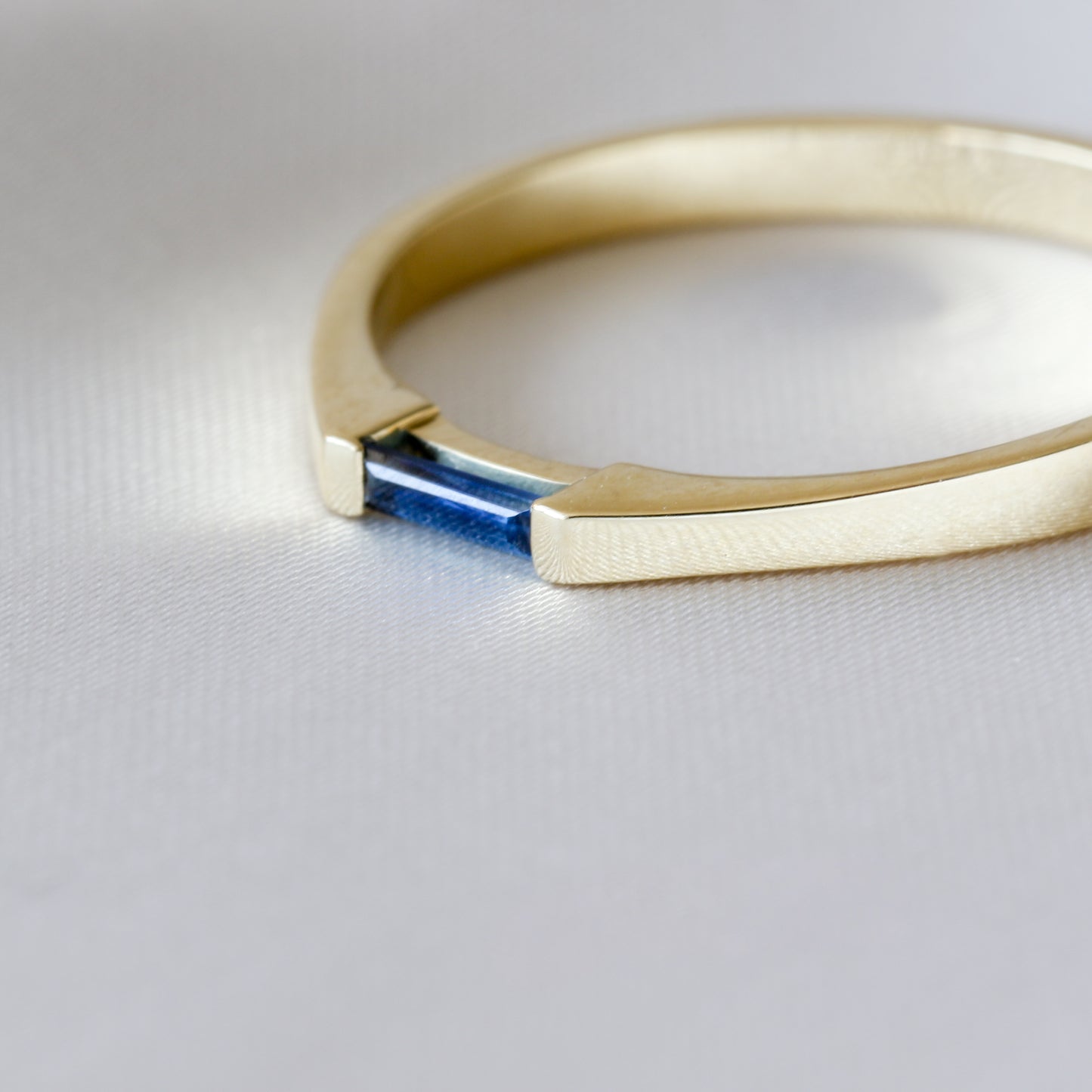 A minimalist style ring with a needle baguette cut blue sapphire set in a 14K yellow gold band, displayed against a white background. close up on stone, ring is in off side view.