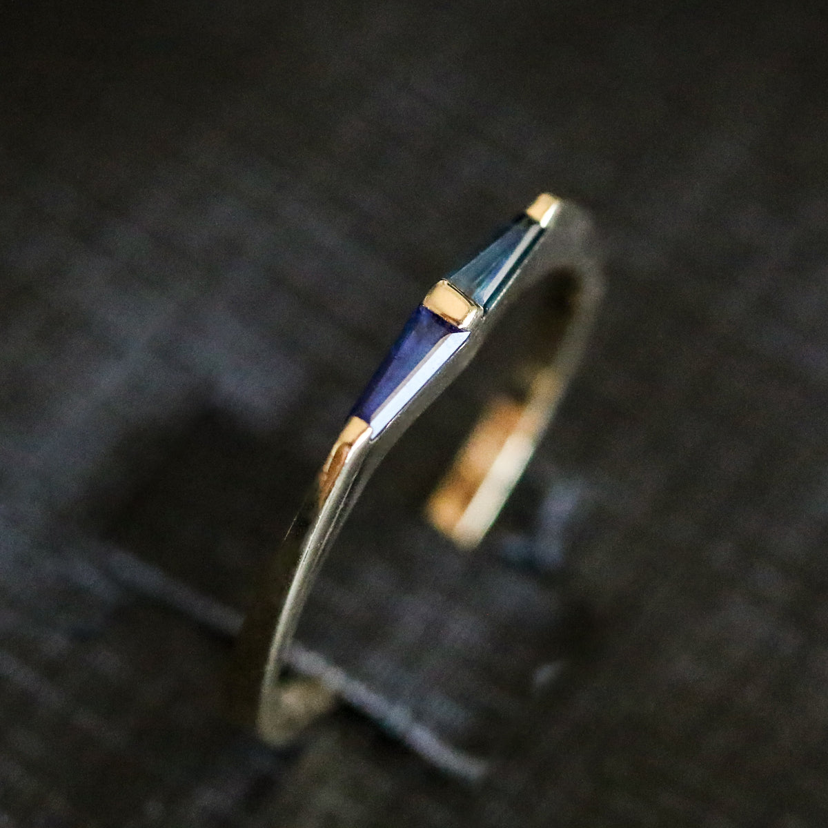 14K yellow gold ring with two tapered baguette blue sapphires and a gold bar between them, on dark fabric