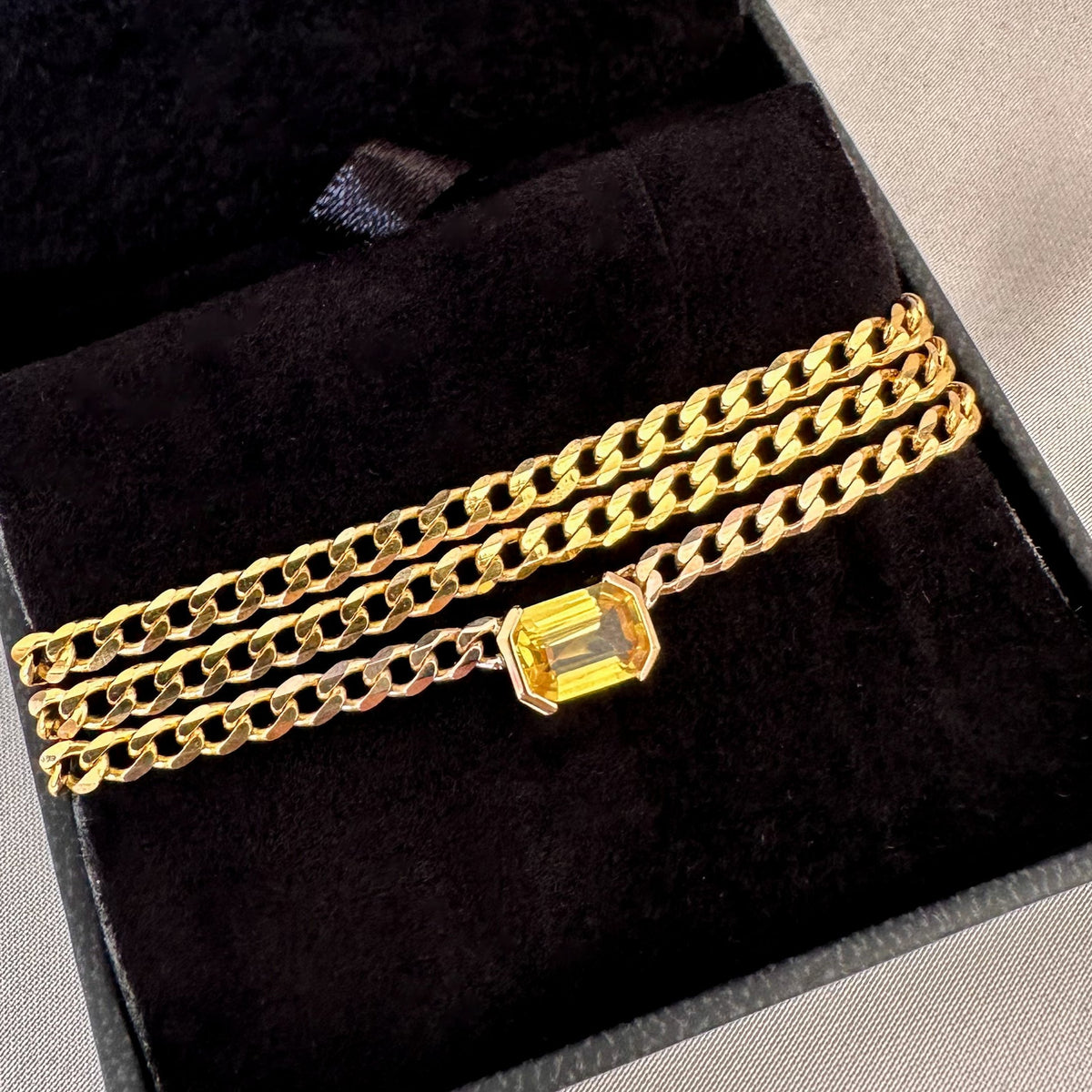Close-up of an east-west emerald cut yellow sapphire in a half bezel on a gold curb chain