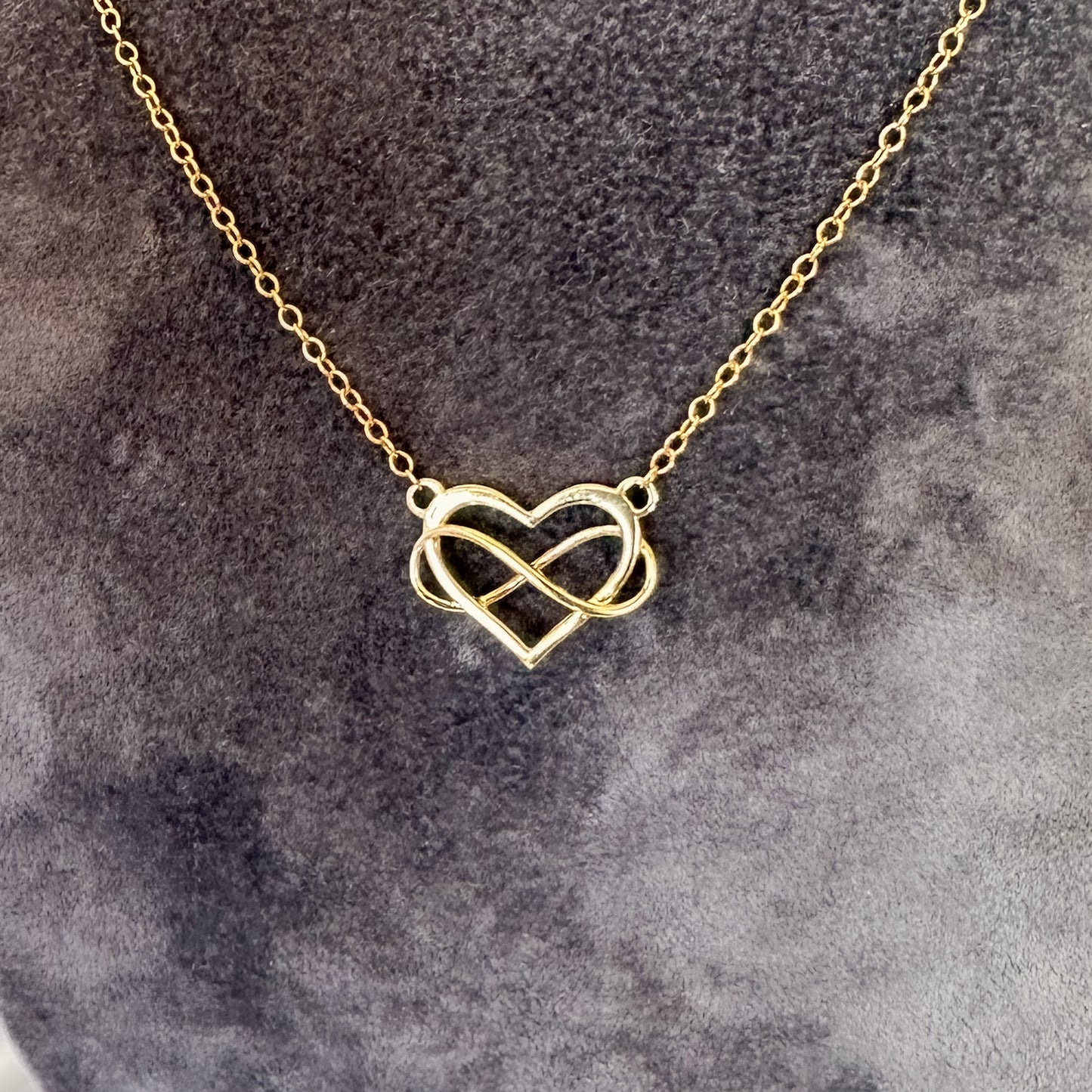 close up yellow gold heart and infinity symbol pendant and chain on gray velvet tourso